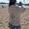 Alexandra, our owner, wearing the Edoaria our mixed cotton embroidered sweatshirt with zipped polo neckline from the Jardin des cyclades collection. Looking at a Ferris Wheel and the sea